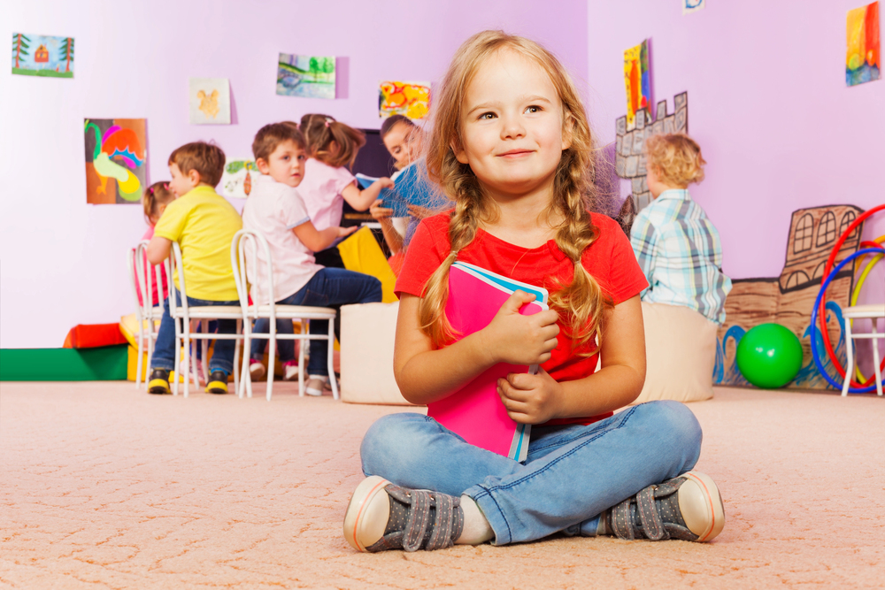 Smiling young girl sitting cross-legged on the classroom floor holding a pink notebook, with other children interacting in the background.